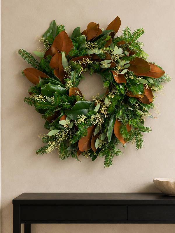 The Evergreen Edit wreath styled on a plaster wall beside a round mirror, with Fraser fir, large magnolia leaves, and seeded eucalyptus