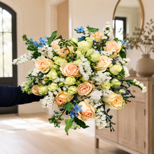 Load image into Gallery viewer, Peach and cream roses with white and blue accents in a hand-held bouquet in a bright entryway
