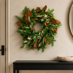 The Evergreen Edit wreath styled on a plaster wall beside a round mirror, with Fraser fir, large magnolia leaves, and seeded eucalyptus
