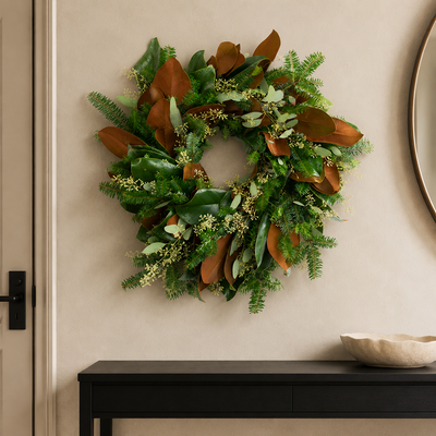 The Evergreen Edit wreath styled on a plaster wall beside a round mirror, with Fraser fir, large magnolia leaves, and seeded eucalyptus