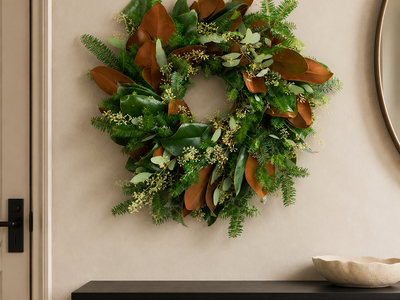 The Evergreen Edit wreath styled on a plaster wall beside a round mirror, with Fraser fir, large magnolia leaves, and seeded eucalyptus