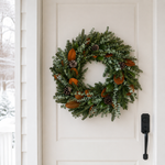 The Welcome Wreath on a white front door in the snow, with Fraser fir, blue-green baby eucalyptus, magnolia, and pinecones